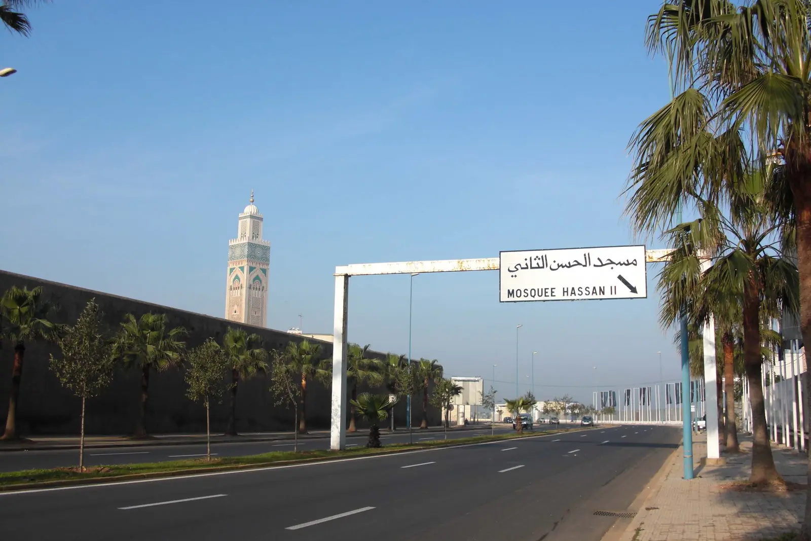 Palm-lined boulevard in Casablanca leading to the minaret of the Hassan II Mosque, with a road sign to the mosque